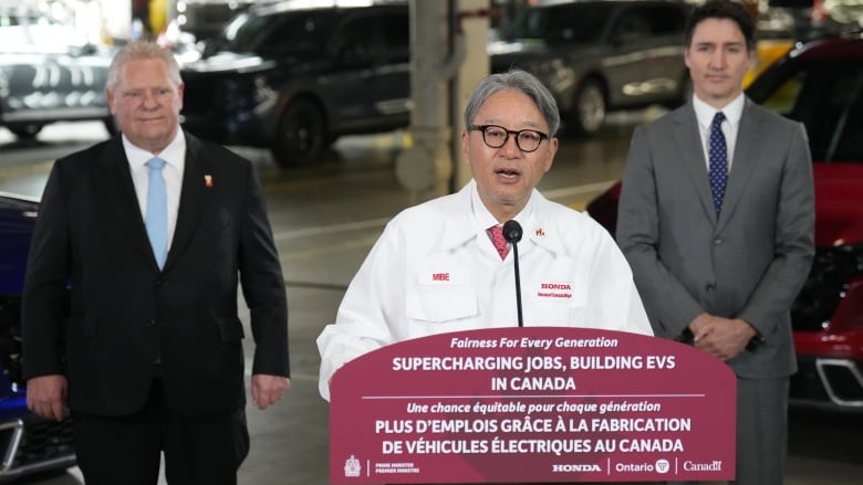 An Asian man speaks at a podium to reporters, while two men stand behind him listening during a news conference at an auto plant.