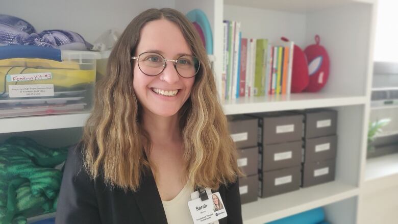 A woman with brown and blonde hair wearing glasses and a black blazer smiles at the camera while standing in front of a white bookcase full of books and other items.