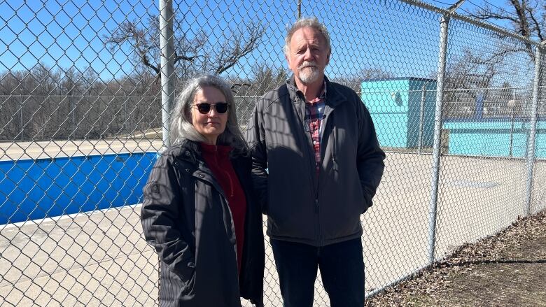 A woman and a man are pictured standing side-by-side in front of a chain-link fence surrounding an empty outdoor pool.