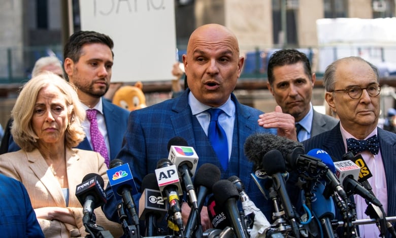 A man in a blue suit and tie stands in front of a dozen news agency microphones in New York City.