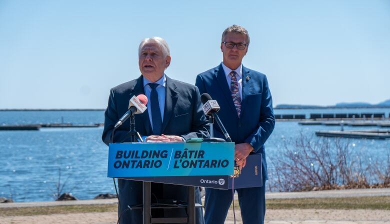 Two men wearing suits stand outside, with a lake behind them.