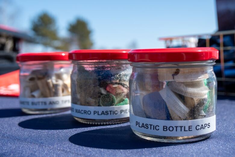 Three glass jars on a table are filled with plastic bottle caps, plastic debris and cigarette butts.