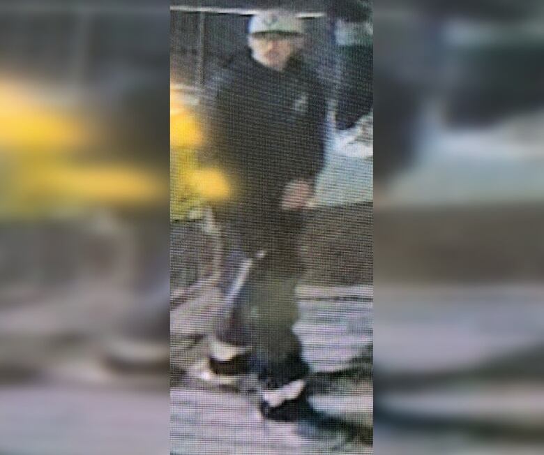 A blurry surveillance video image of a man. He is dressed in black with a grey baseball cap.