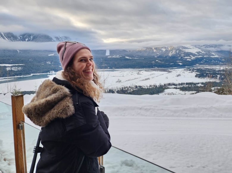 A smiling woman wearing a parka and pink toque poses for a outdoors photo in the winter. A mountain range is pictured in the background.