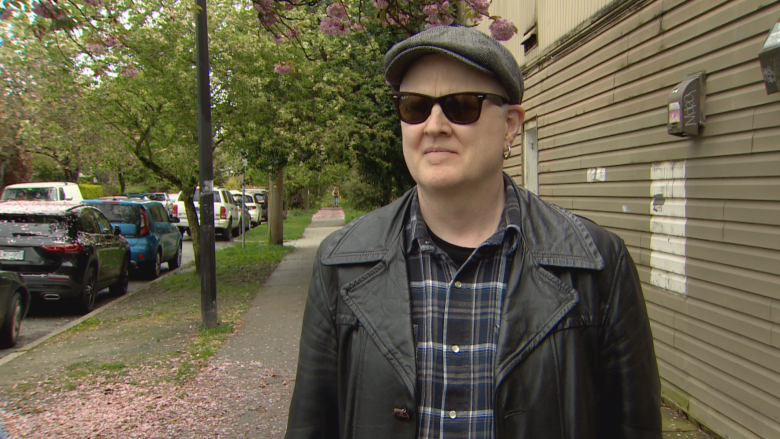 A man in sunglasses and a black jacket stands outside a building.