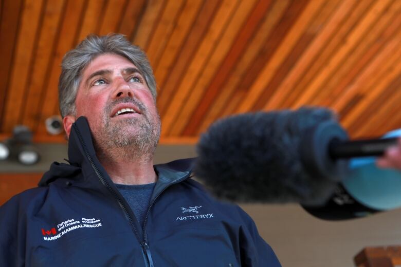 A white man wearing a blue jacket speaks to a pair of mics.