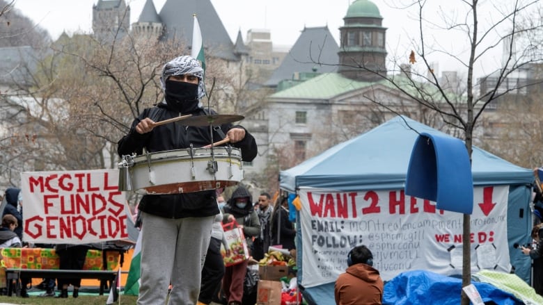 Wearing a keffiyeh, a person plays a portable drum at an encampment, surrounded by other pro-Palestinian protesters at McGill University. 