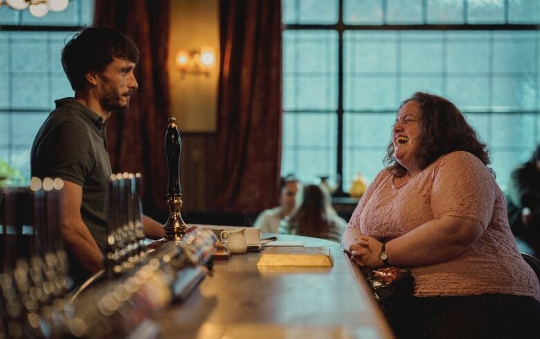 A man stands behind a bar, looking at a woman laughing while sitting at the bar.