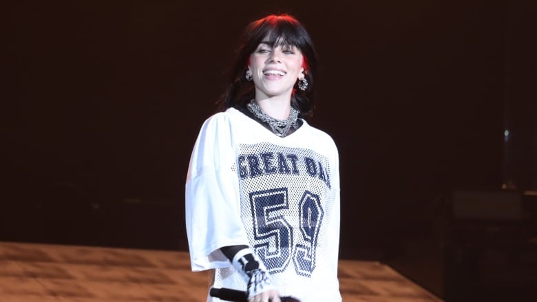 A smiling woman wearing a sports jersey and holding a microphone stands on a stage. 