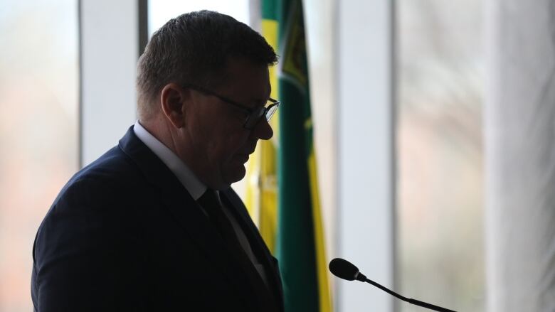 A man in a dark suit and a white shirt stands in front of a podium. The Saskatchewan flag is in the background. 