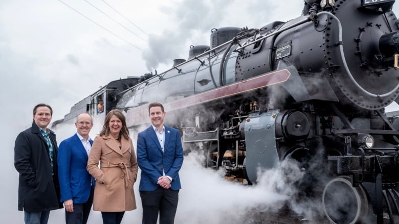 Four people stand in front of a locomotive train engine, puffing with steam.