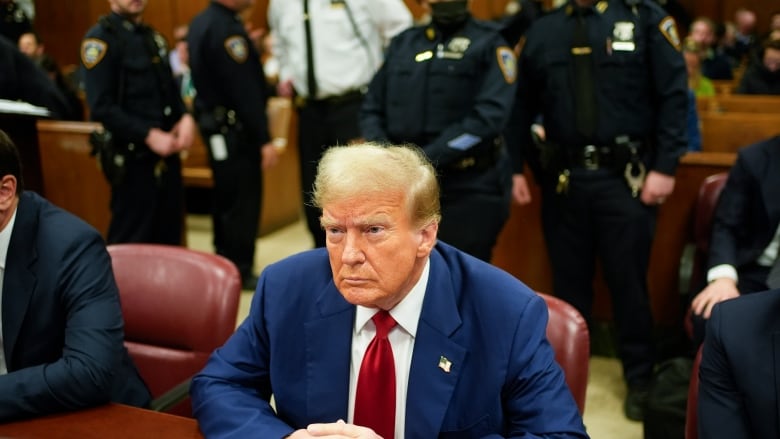 Trump, in a blue suit and red tie sits with his hands on the table in front of him. Several police officers are behind him in the courtroom.