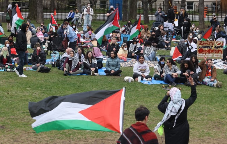 People sitting or standing on a lawn at Uniersity of Ottawa, listening to a woman speaking with a mic and a man standing next to her holding a Palestinian flag.