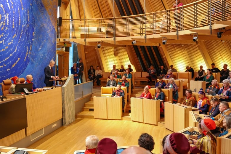 People sit behind podiums in a large light wood auditorium.