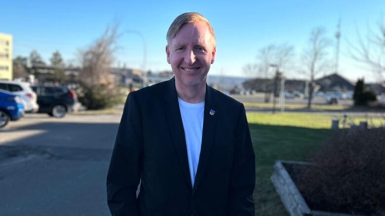 Man in dark suit jack with white collarless shirt underneath, standing outside and smiling directly at the camera. 