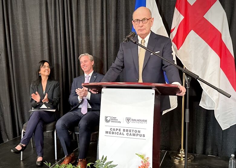 A man in a blue suit with a white shirt, yellow tie and glasses stands at a podium with two flags behind him while another man and a woman applaud.