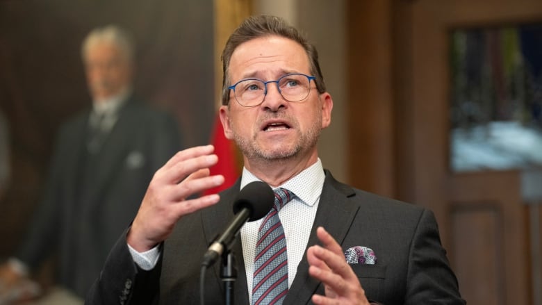 A man in a dark suit and striped tie speaks into a microphone. A portrait hangs in the background.