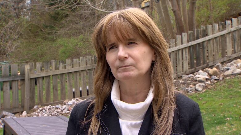 A woman stands outside in front of a fence. She has light brown hair, wears a white turtle neck and a black blazer. 