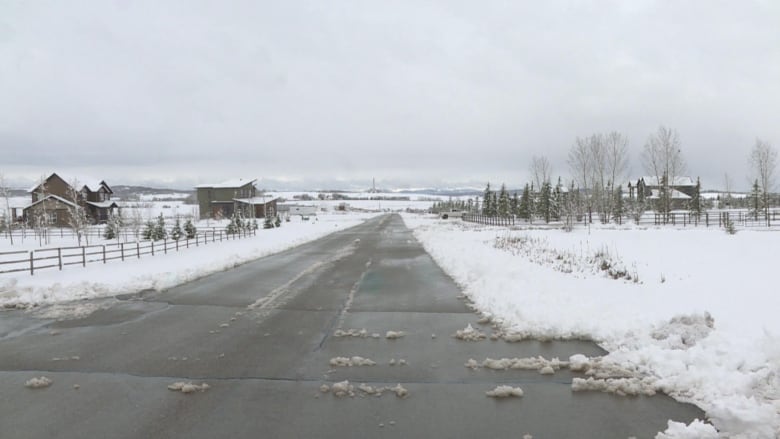 a road surrounded by large homes. there is snow on the side of the road, and a mountain range is pictured in the background.