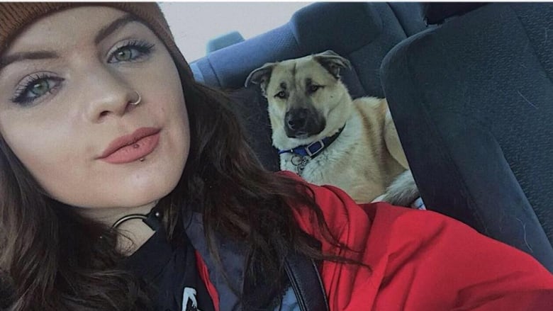 A selfie of a young woman with dark hair, sitting in a car, with a dog in the back seat behind her.