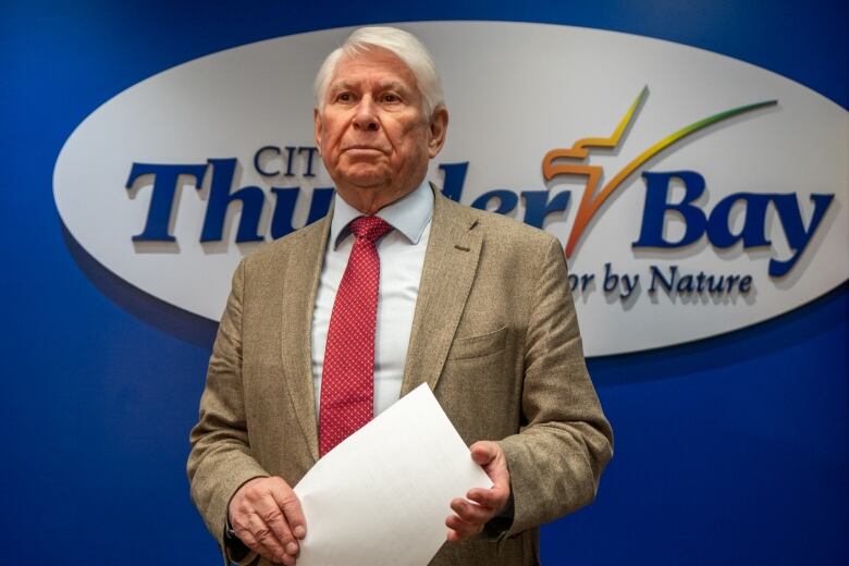 A man wearing a suit stands in front of a blue wall that says 