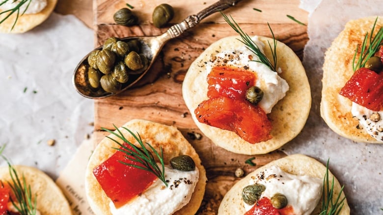 Closeup overhead shot of Mini Blinis topped with Smoked Tomato, cream cheese, dill and capers. They're sitting on a wooden cutting board with a spoon of capers next to it. 