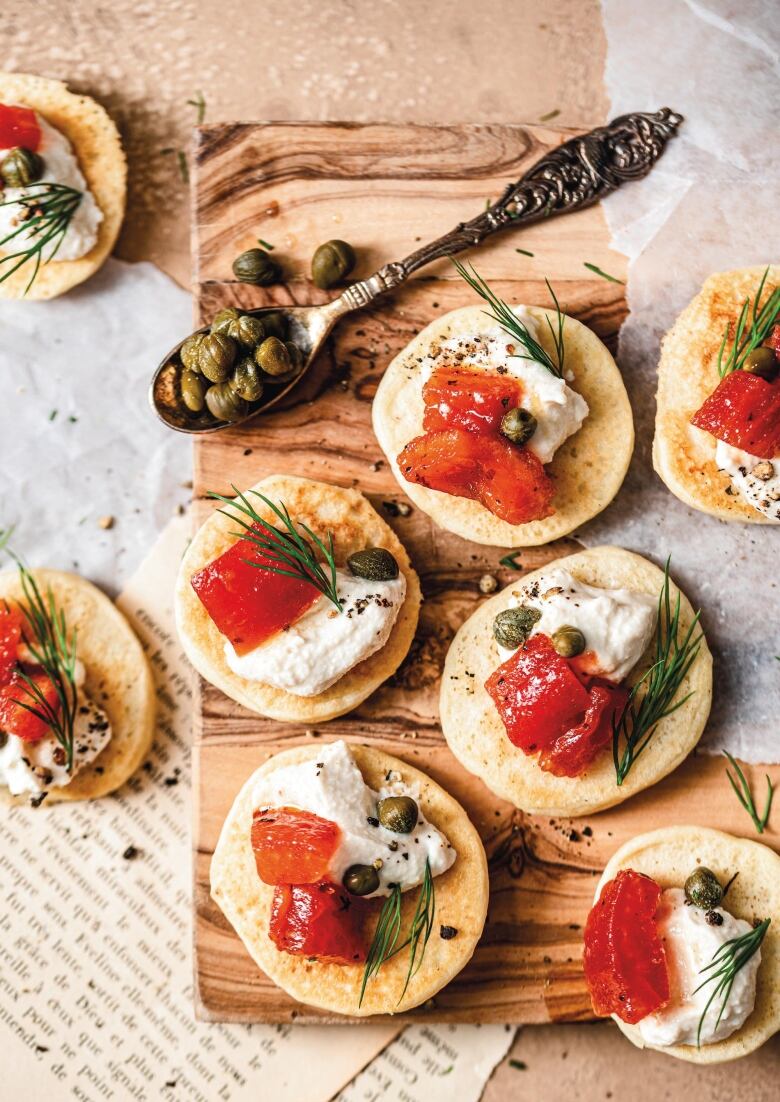 Overhead shot of Mini Blinis topped with Smoked Tomato, cream cheese, dill and capers. They're sitting on a wooden cutting board with a spoon of capers next to it.