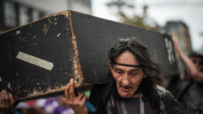 A weathered-looking man carries a fake wooden coffin.