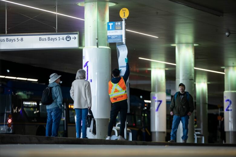 A man in an orange high-vis vest changes out a bus schedule at a stop.