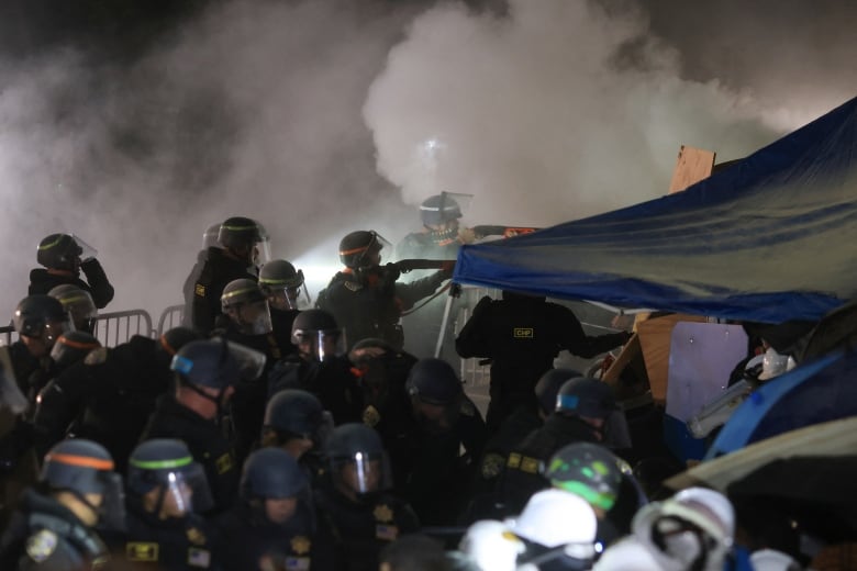 Several helmeted police officers are shown near a canvas enclosure, with large amounts of smoke shown in the background.