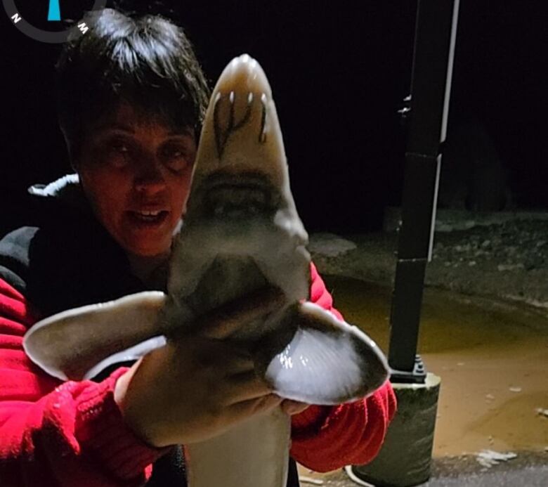 Standing on shore of a river, a woman holds a large fish up to the camera.