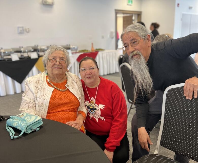 A woman wearing a red sweater crouches down next to a woman sitting at a table. A man with a long, grey beard stands next to them.