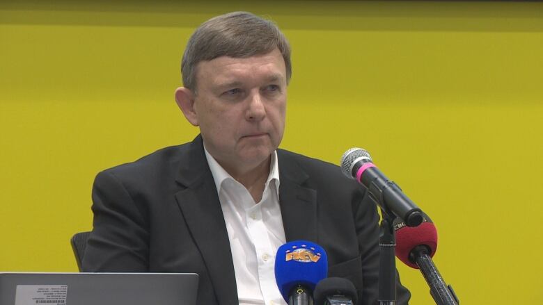 A man wearing a suit sits in front of microphones. He sits against a yellow wall.