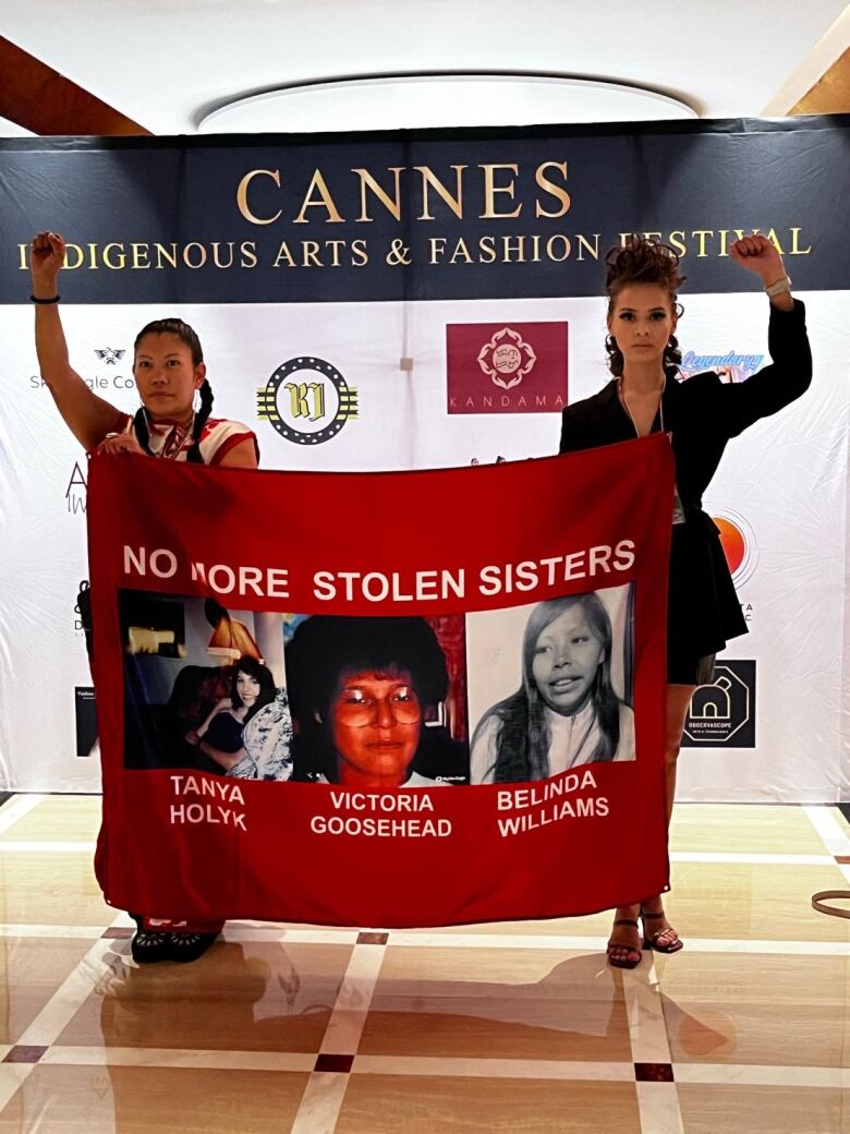 Two women hold up a banner with three women's faces and names on it saying 