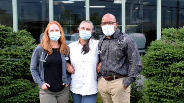 Three people wearing medical masks stand together on a lawn, with windows to a medical clinic in the background. 