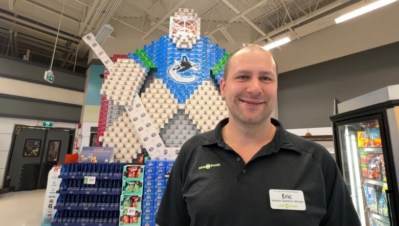 A shot of a smiling man at a grocery store. There's a giant sculpture of a hockey player made using boxes in the background.