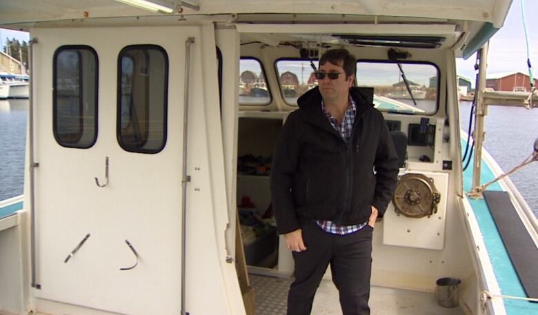 Man with brown hair, a jacket and checked shirt stands on a fishing boat and wears sunglasses. 