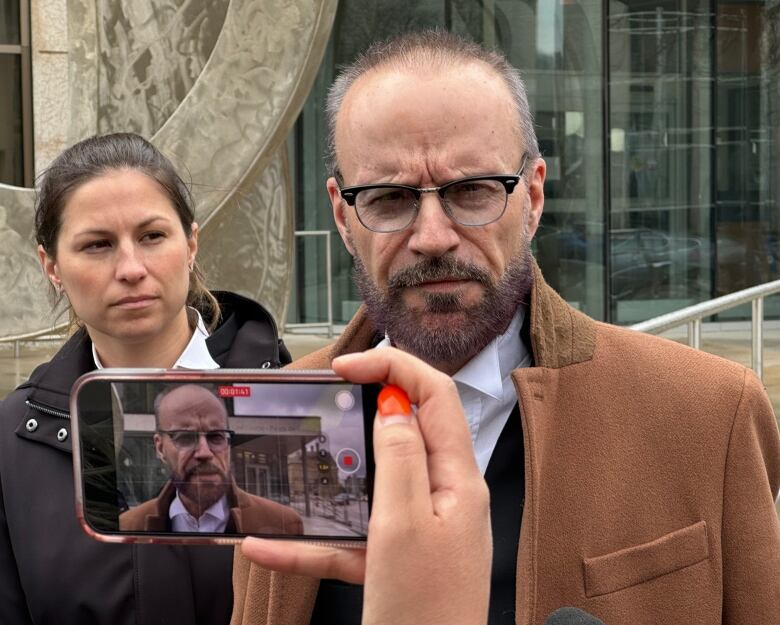 A man and a woman stand outside a courthouse. A person is recording video of the man on a cellphone.