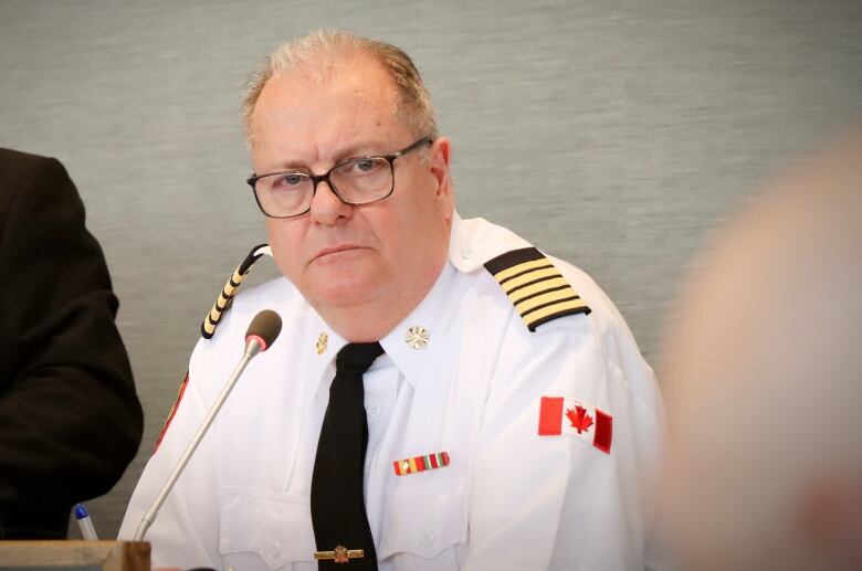 A man with thinning hair and glasses wearing a white uniform shirt with black and gold epaulettes and a black tie sits at a microphone and listens.