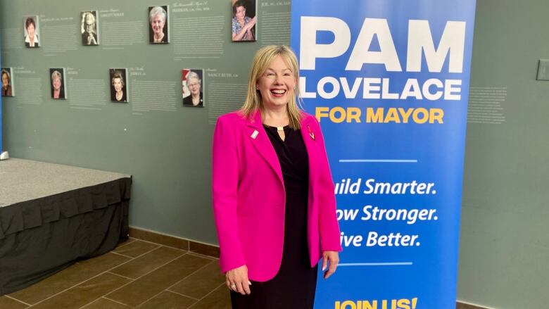 A woman wears a black shirt with a pink blazer in a grey room.