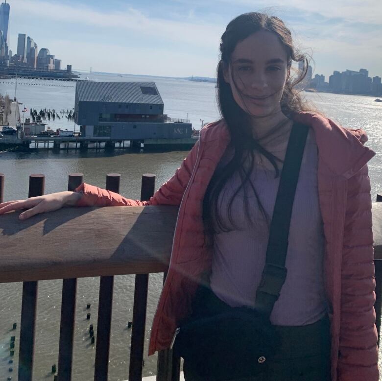 Mina Jordanides leaning against a wood post wearing a pink jacket, purple shirt, black pants and has a Lululemon bag around her body. 