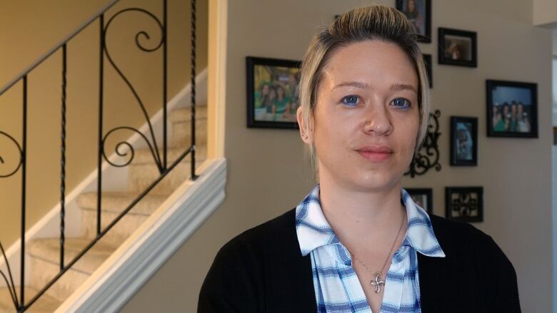 A woman with her blond hair up stares at the camera with a serious expression. She's inside a home and behind her family portraits can be seen covering the wall.