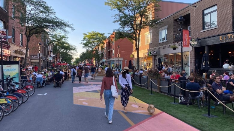 Two people walk a dog down a street closed off to traffic. Others are walking around up ahead and people are sitting at dining tables on the sides.