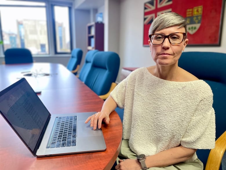 Woman with short hair and glasses sits at a boardroom table infront of a laptop.