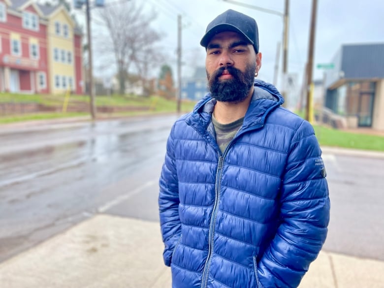 Man with blue jacket, hat, and beard stands in rain on street corner.