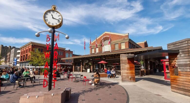 A photo of a large brick building. There are pedestrians sitting outside at public tables and a large clock with the words 'ByWard Market' written along its sides.