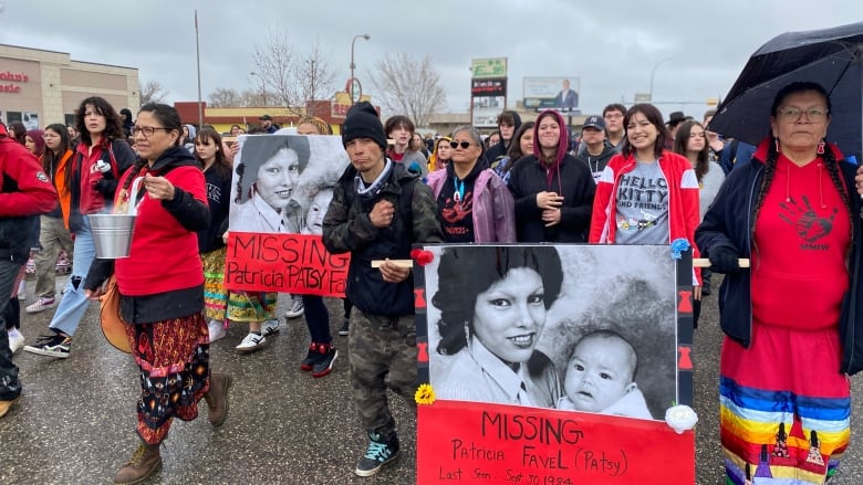 A group of people, wearing bright and dark collaborative colors walk together down a street, while carrying a enlarged photo of a woman.