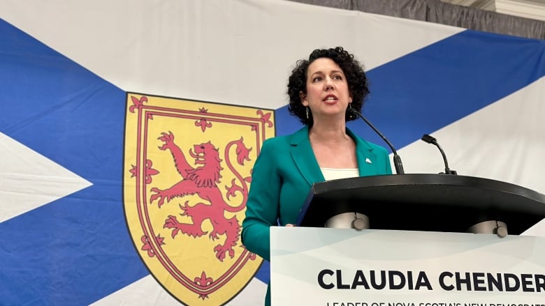 A woman in a green suit speaks at a podium in front of a large Nova Scotia flag.