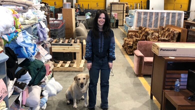 A woman with her dog stands in a warehouse surrounded by clothes and furniture