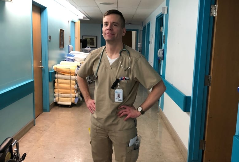 A man wearing scrubs stands in a hospital hallway with a stethoscope hanging around his neck. 